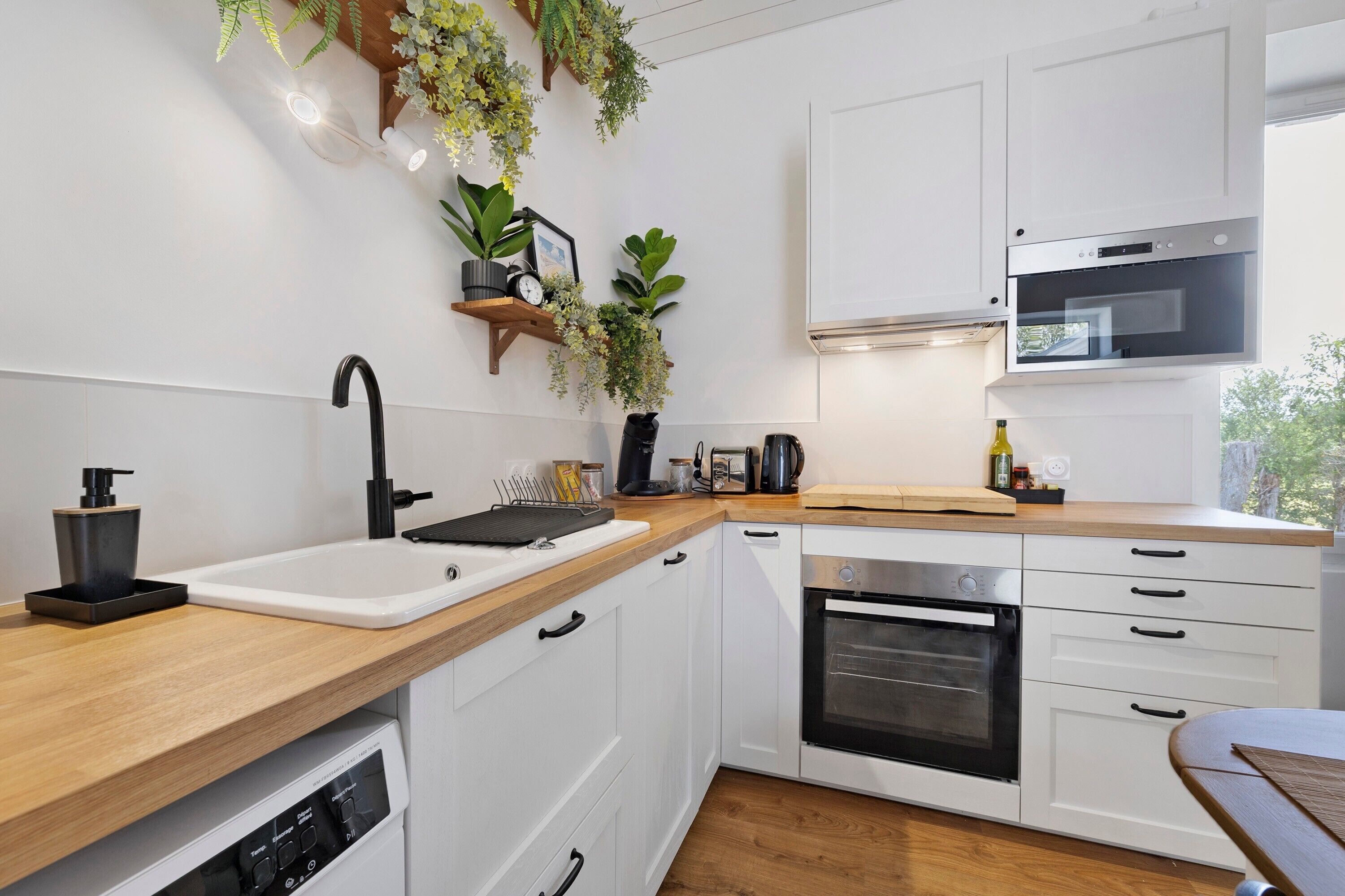 A modern kitchen with white cabinetry, a wooden countertop, a black faucet, an oven, and hanging plants.