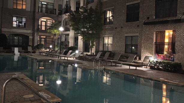 Une zone de piscine éclairée pour la baignade nocturne, avec des chaises longues, et un bâtiment doté de nombreux balcons en arrière-plan.