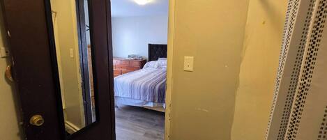 Uma porta dá acesso a um quarto com cama, cômoda e luminária no teto.