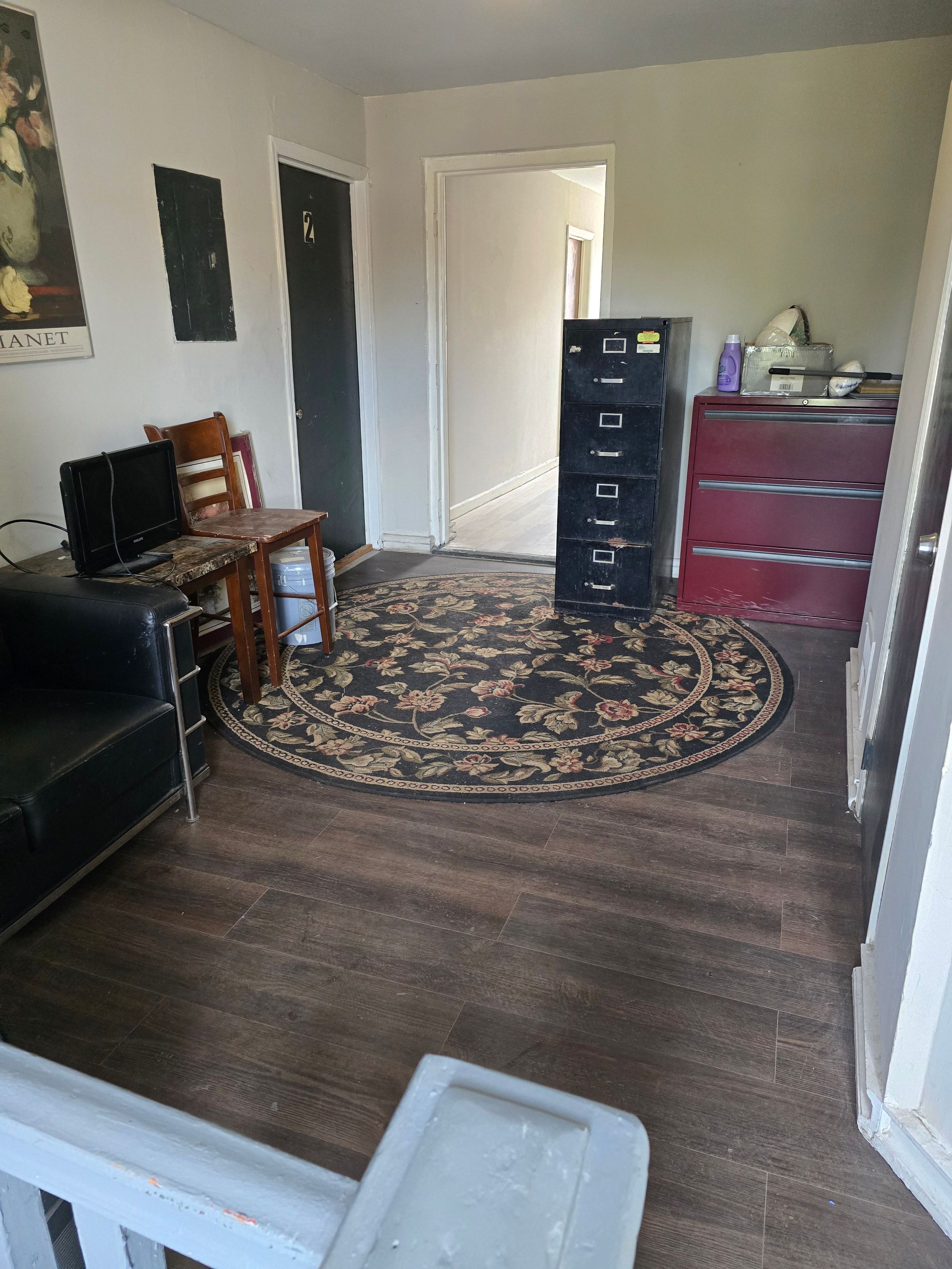 A room with a dark wood floor, a floral rug, a black sofa, a wooden chair, a desk with a computer, and a red filing cabinet.