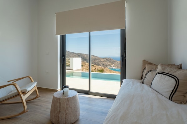 Ein Schlafzimmer mit einem Bett, einem Schaukelstuhl, einem runden Holztisch und einer großen Schiebeglastür, die zu einem Pool und einer schönen Aussicht führt.
