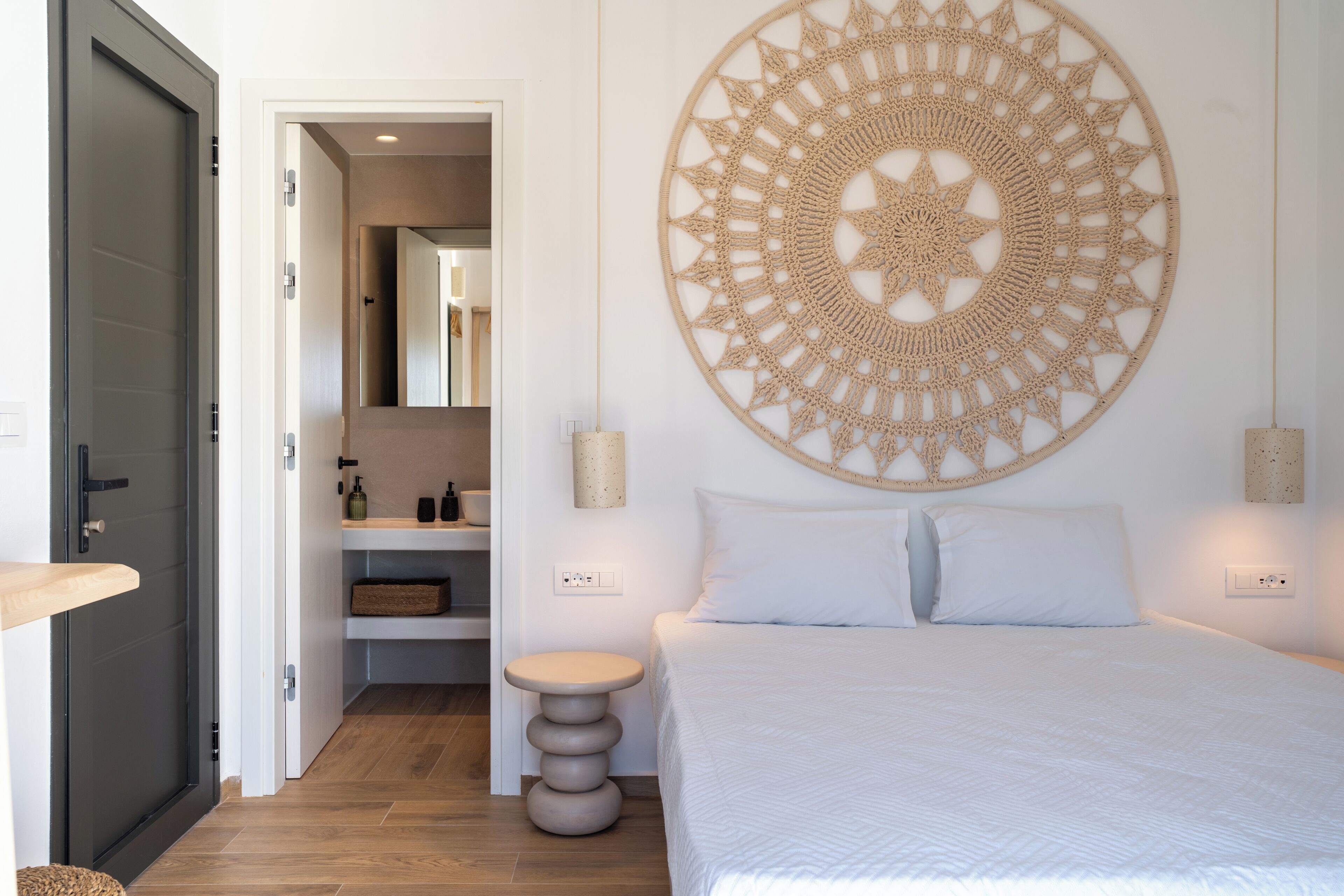 Um quarto com uma cama, um enfeite circular na parede, uma mesa de cabeceira e uma porta para o banheiro.