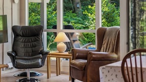 Une pièce avec une grande fenêtre, un fauteuil en cuir noir, un fauteuil en cuir marron, une petite table avec une lampe et une chaise en bois.