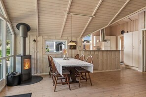 A cozy room with a wood-burning stove, a dining table with chairs, and a kitchen area with cabinets and a window. - Holiday home at Kvie Sø (Ansager)