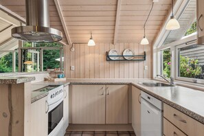 A kitchen with wooden cabinets, a stainless steel range hood, a dishwasher, and a window with a view of greenery. - Holiday home at Kvie Sø (Ansager)