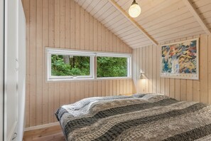 A bedroom with wooden walls, a large window, a bed with a striped blanket, and a painting on the wall. - Holiday home at Kvie Sø (Ansager)