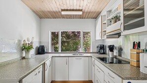 Una cocina con armarios blancos, techo de madera, ventana con vista a árboles y encimera con cafetera y utensilios.