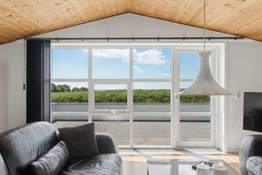 A modern living room with a large sliding glass door, a black leather sofa, a coffee table with candles, and a hanging pendant light. - Spacious holiday home with water view (Redsted)