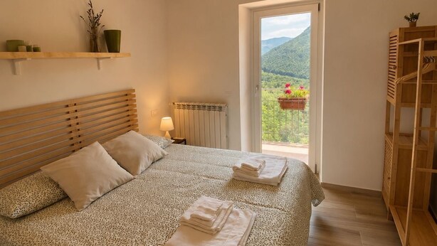 A bedroom with a bed, a radiator, a window with a view, and a wooden shelf.
