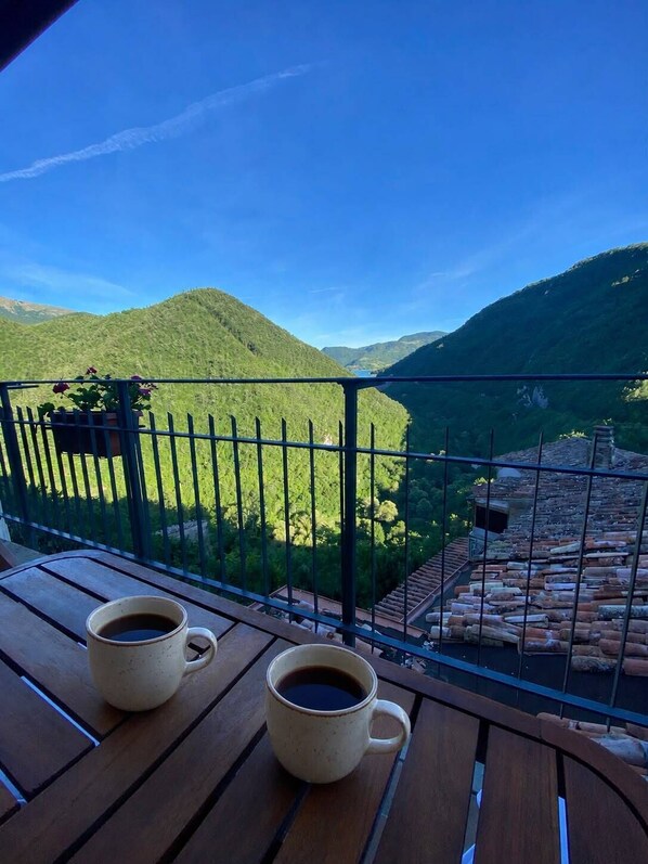 A balcony with two coffee cups, a metal railing, and a view of green hills and a river.
