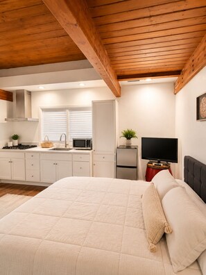 A bedroom with a bed, a television, a small kitchen area, and a wooden ceiling.