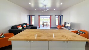 A modern living room with a central wooden table, a sofa with colorful cushions, an orange chair, a television, and a ceiling fan. - Kuki Kainga (Matavera District)