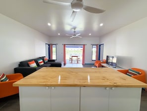 Una sala de estar moderna con una mesa de madera en el centro, un sofá con cojines coloridos, un sillón naranja, un televisor y un ventilador de techo.