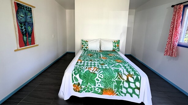 A bedroom with a bed, a ceiling fan, a window with curtains, and a wall painting. - Kuki Kainga (Matavera District)