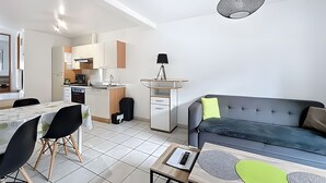 Ein modernes Wohnzimmer mit grauem Sofa, einem Esstisch mit Stühlen, einer kleinen Küchenzeile und einer Deckenleuchte.
