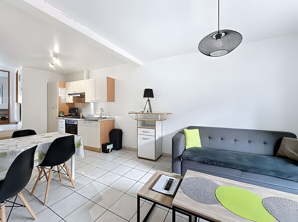 A modern living room with a grey sofa, a dining table with chairs, a small kitchen area, and a ceiling light fixture.