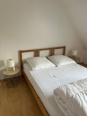 A single bed with a wooden headboard, two white pillows, and a white duvet. There are two bedside tables with white lamps and a roll of toilet paper on one. The room has wooden flooring and white walls.