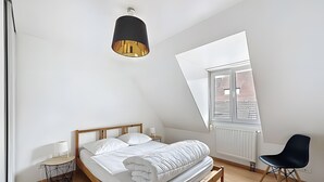 Ein Schlafzimmer mit schrägen Wänden, einem Bett mit weißer Bettwäsche, einem schwarzen Stuhl, einem Nachttisch und einer besonderen Deckenleuchte.