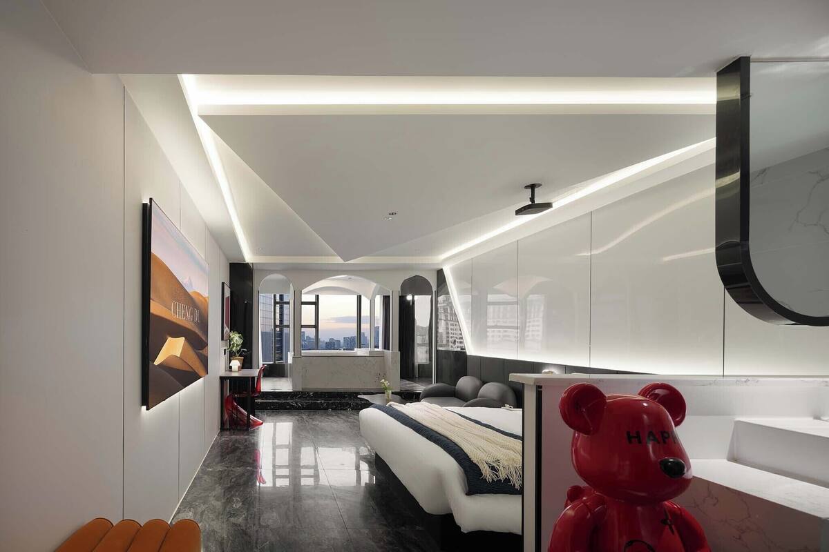 Une chambre d’hôtel moderne avec un lit, un ours en peluche rouge, une vue sur la ville et un design minimaliste.
