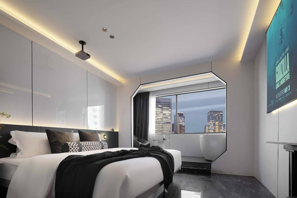 Une chambre d’hôtel moderne dotée d’un grand lit, offrant une vue sur la ville, d’un écran de projection et d’un design minimaliste.