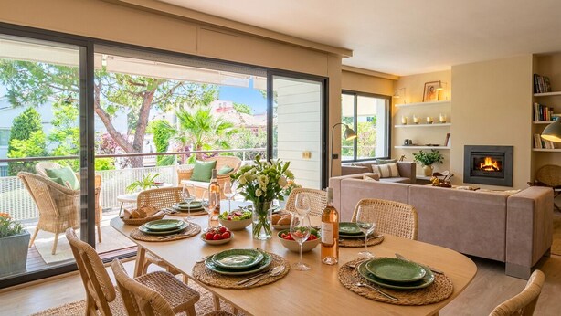 Una zona pranzo con tavolo imbandito per quattro persone, un divano, un camino e un balcone con mobili in vimini e piante.