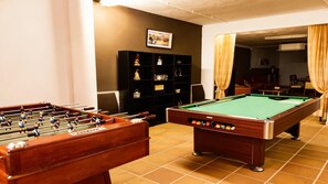 Een biljart tafel, een tafelvoetbalspel en een plank met diverse spullen in een kamer met betegelde vloer.