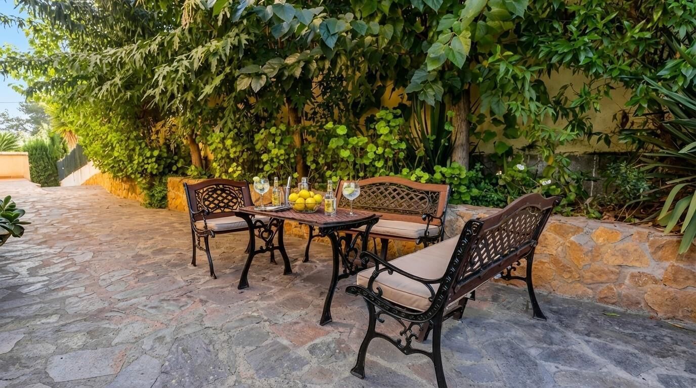 Een terras buiten met stenen vloer, een houten tafel, twee stoelen met kussens, en een bank.
