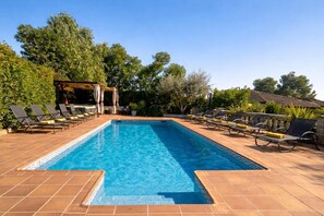Ein rechteckiger Swimmingpool mit gefliesten Rändern, umgeben von Liegestühlen und einer Pergola mit Vorhängen.