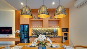 A dining area with a wooden table set for four, overhead pendant lights, a TV on the wall, and a kitchen in the background. - Villa Omara - Nusa Dua (Nusa Dua)