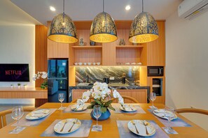 Un comedor con una mesa de madera preparada para cuatro personas, lámparas colgantes en el techo, un televisor en la pared y una cocina al fondo.