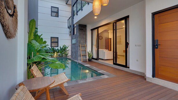 A modern outdoor pool area with wooden decking, a table with chairs, and a large sliding glass door leading to a bright interior. - Villa Omara - Nusa Dua (Nusa Dua)