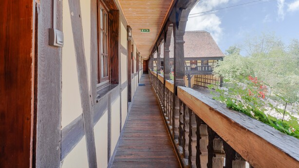 A corridor with wooden railings and a wooden ceiling, leading to a building with a tiled roof. - Gite Le Coquelicot "bi's anstetts" – Authentic Alsatian Farmhouse Retreat (Issenhausen)