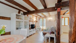 A cozy room with exposed wooden beams, a white cabinet with glass doors, a television, a dining table with a patterned tablecloth, and a window with a view. - Gite Le Coquelicot "bi's anstetts" – Authentic Alsatian Farmhouse Retreat (Issenhausen)
