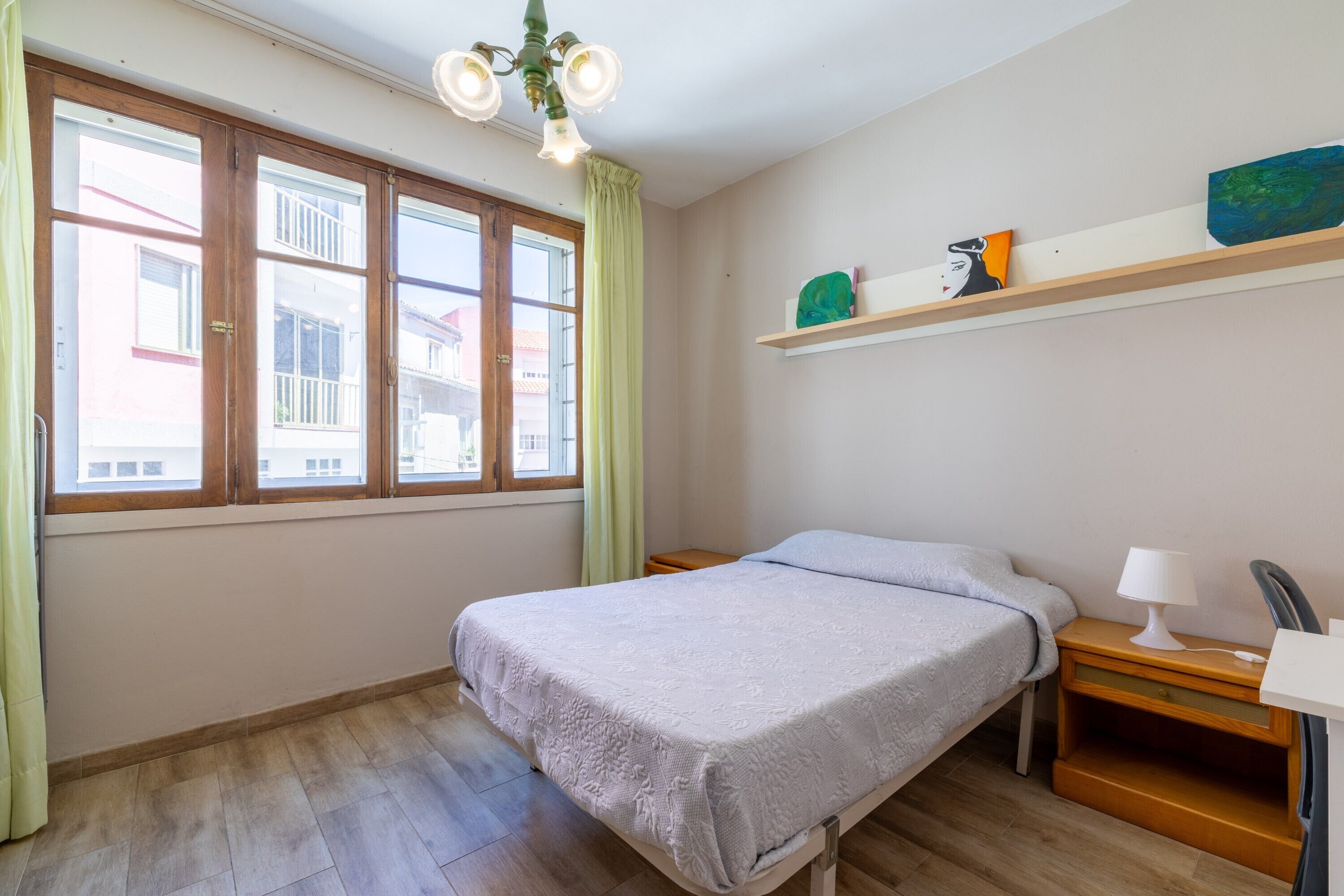 A bedroom with a bed, a nightstand, a lamp, a shelf with decorative items, and a window with curtains.