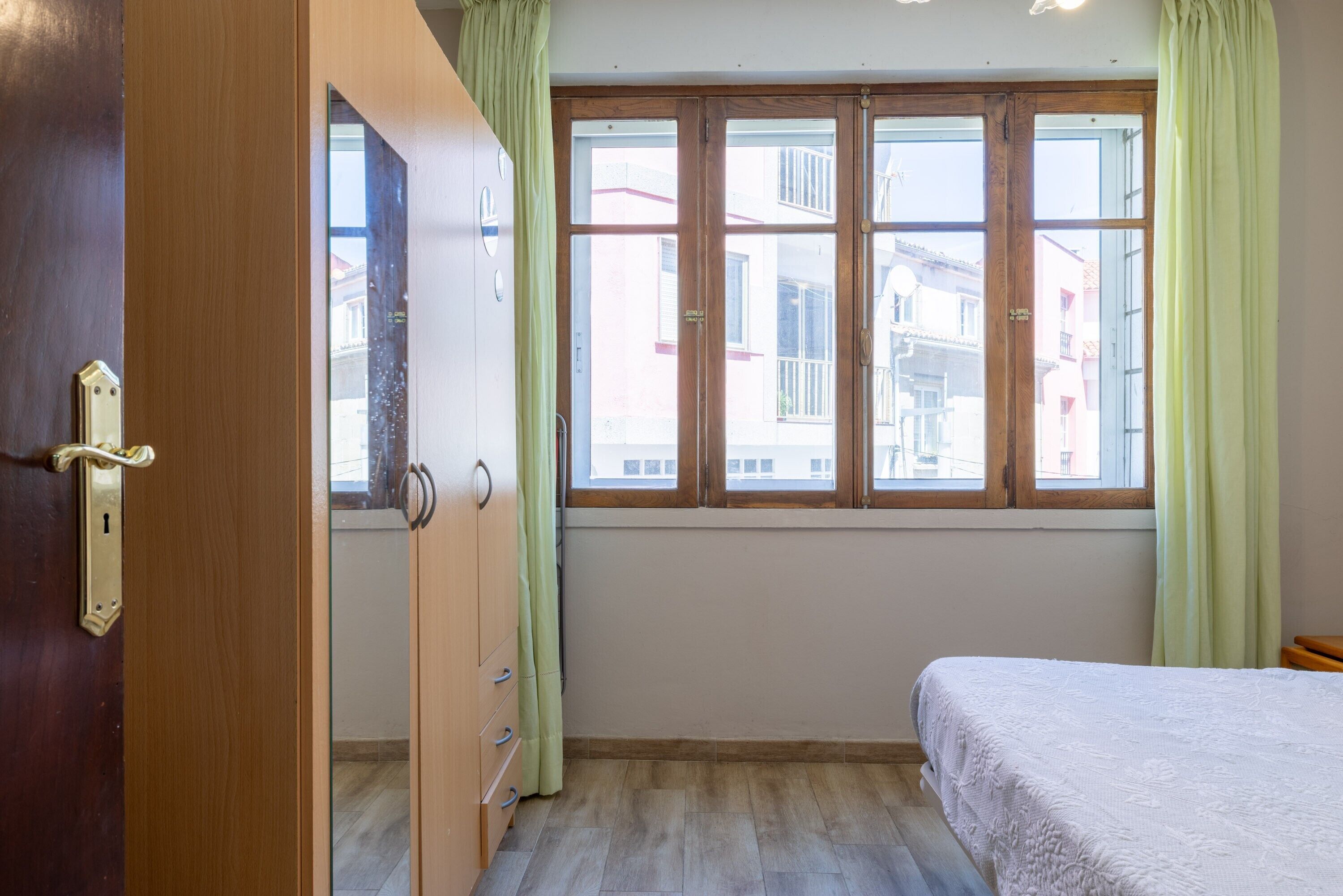 Une pièce avec un lit, une armoire en bois, une fenêtre avec des rideaux et une vue sur les bâtiments à l’extérieur.