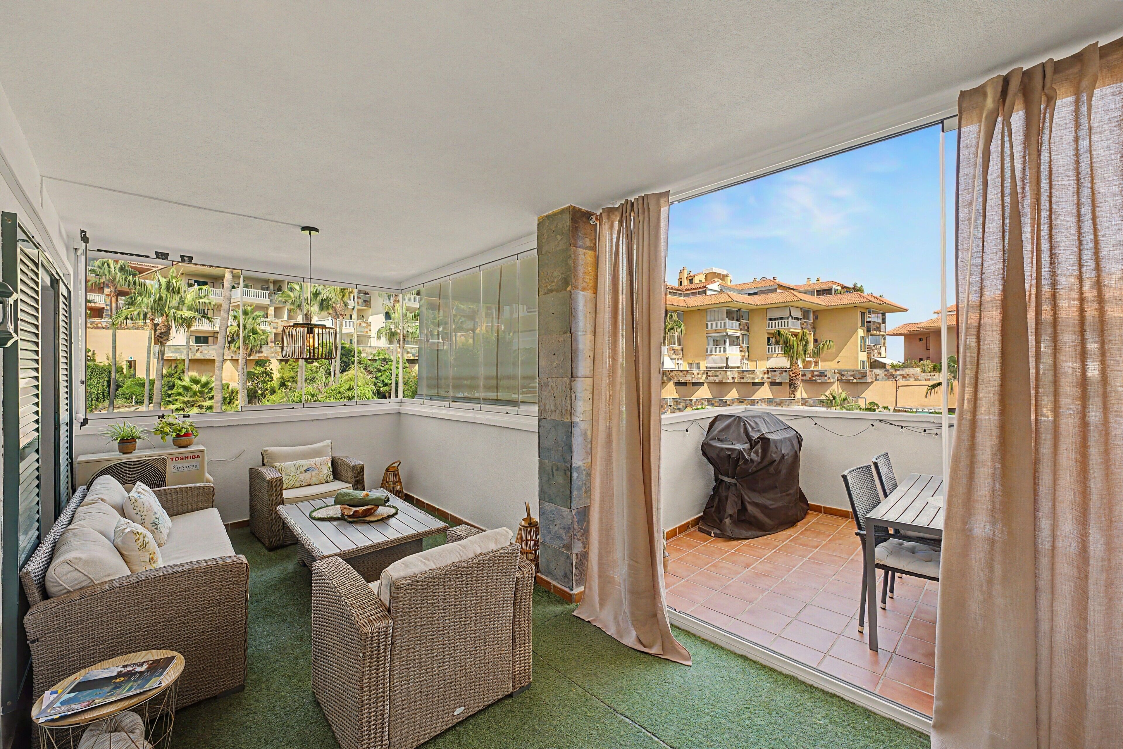 A balcony with wicker furniture, a grill, and a view of a residential area.