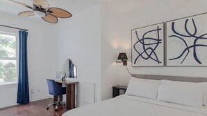 Een slaapkamer met een bed, een bureau met stoel, een plafondventilator, kunst aan de muur en een raam met gordijnen.