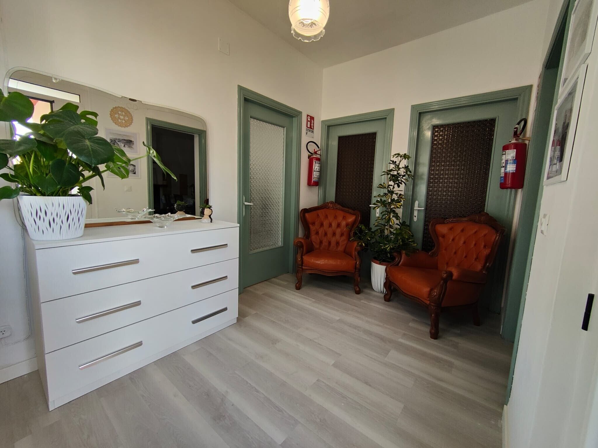 Un couloir avec une commode blanche, deux chaises en bois, une plante en pot et un extincteur.