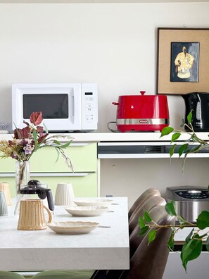 Eine moderne Küche mit einer weißen Mikrowelle, einem roten Slow Cooker und einer Kaffeemaschine. Auf der Arbeitsfläche steht eine Vase mit Blumen und an der Wand hängt ein gerahmtes Bild.