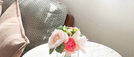 A round marble table with a clear glass vase containing flowers, positioned next to a grey patterned sofa with a wooden armrest.