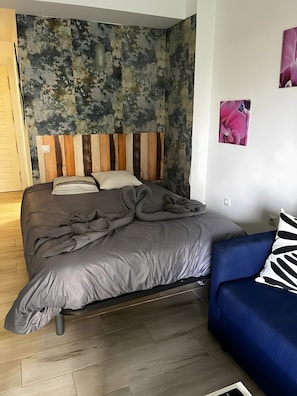 Ein Schlafzimmer mit einem Bett, einem blauen Sofa, einem Holz-Kopfteil und einer Wand mit einem strukturierten Wandbild.
