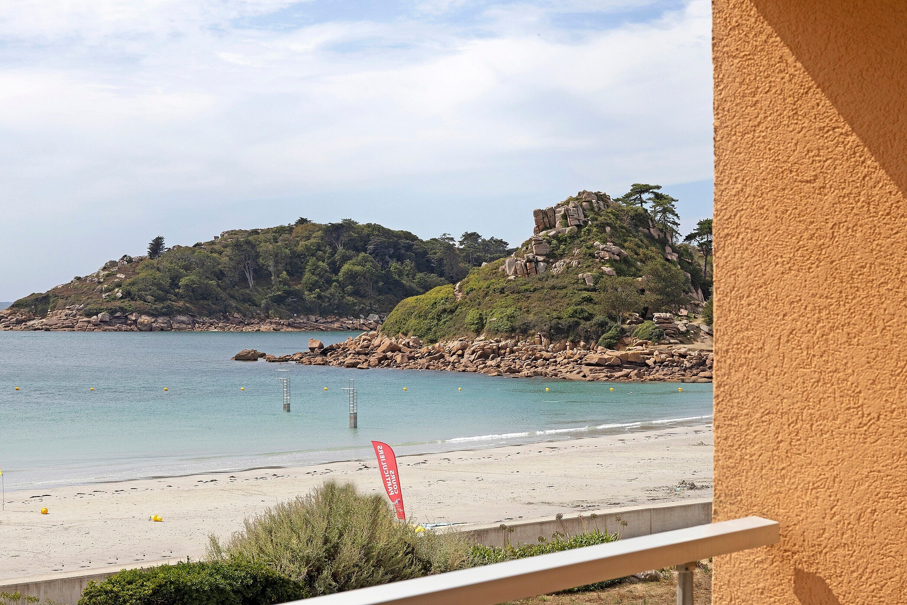 A beach with clear water, a rocky hill in the background, and a red sign.