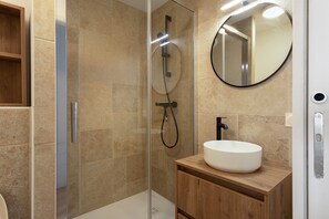 A modern bathroom with a glass-enclosed shower, a wooden vanity with a white sink, and a round mirror.