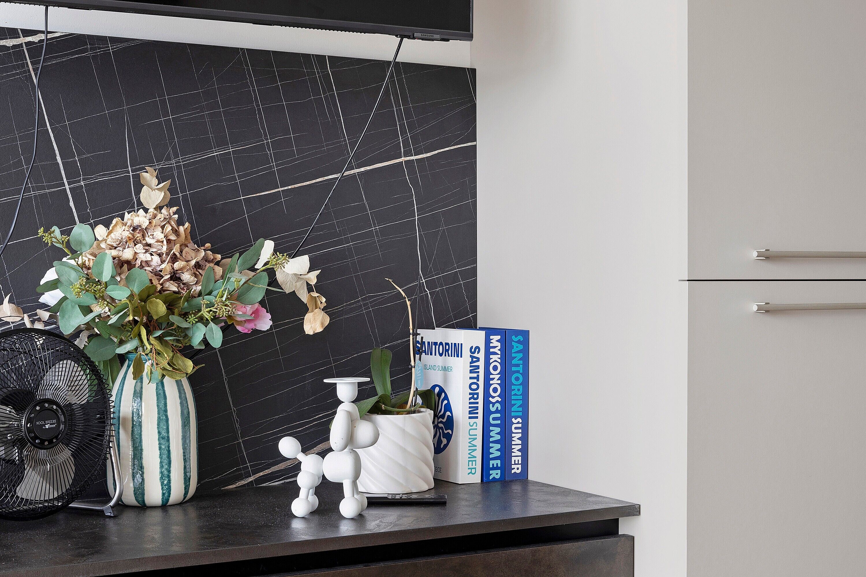 A modern kitchen with a black marble backsplash, a white vase with dried flowers, a small white sculpture, and a fan.