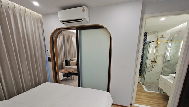 Ein Hotelzimmer mit einem Bett, einem Schreibtisch, einer Klimaanlage, einem Badezimmer mit Dusche und einer Glastür, die zu einem Sitzbereich führt.