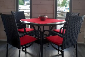 Ett runt bord med en röd duk, omgivet av fyra svarta flätade stolar med röda kuddar, placerat framför en vägg av trä med utsikt över båtar genom ett fönster.