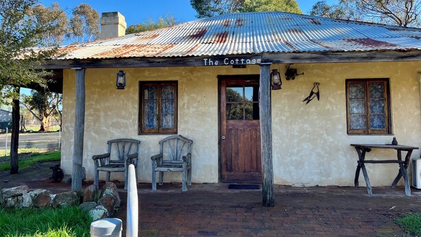 Una construcción rústica con el rótulo 'The Cottage', techo de metal corrugado, dos puertas de madera y una pequeña mesa con una silla afuera.