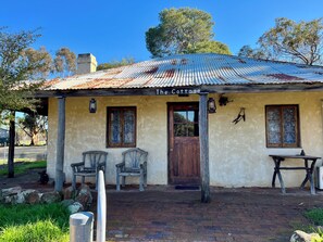 Una construcción rústica con el rótulo 'The Cottage', techo de metal corrugado, dos puertas de madera y una pequeña mesa con una silla afuera.