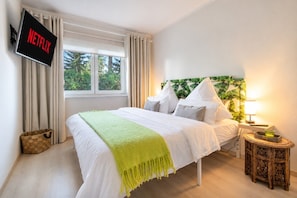 A hotel room with a bed, a TV mounted on the wall, a window with curtains, and a bedside table with a lamp. - Apartment 'Lakeside Retreat Apartment – Kiefernwald-Spaziergänge zwischen Berlin und Tropical Islands' (Bestensee)