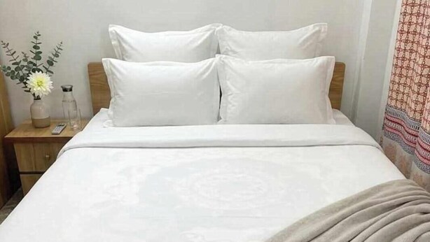 Uma cama com roupa de cama branca, cabeceira de madeira, mesa de cabeceira com um vaso e uma garrafa, e cortina estampada.